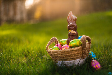 Easter eggs in basket with easter bunny on top. Chocolate rabbit with colorful decorated eggs in wicker basket in grass. Magical morning light, spring season holidays. Traditional egg hunt