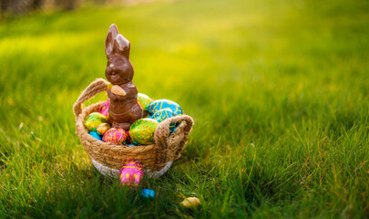 Easter eggs in basket with easter bunny on top. Chocolate rabbit with colorful decorated eggs in wicker basket in grass. Magical morning light, spring season holidays. Traditional egg hunt