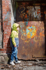 A little boy in a yellow jacket and a hoodie, by an old abandoned wall in a city park, is interested in graffiti