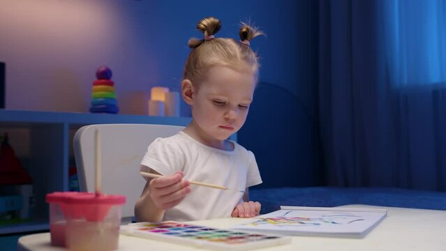 Cute two-year-old blonde girl paints multicolored paints on white paper sitting at home at table in evening.