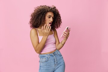Woman blogger holding phone with curly hair in pink top and jeans poses on pink background, copy space, technology and social media