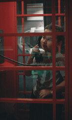Obraz na płótnie Canvas In a London red telephone booth, a child boy is holding a phone while talking to his mother. Rebeok calls home from an old red telephone booth. Concept of a child in a retro telephone booth.