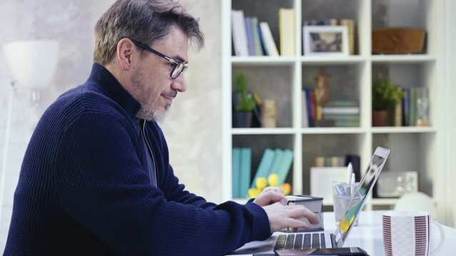 Businessman typing on with laptop computer. Home office, working online from home, browsing internet. Portrait of mature age, middle age, mid adult man in 50s. Bearded, gray hair, glasses.