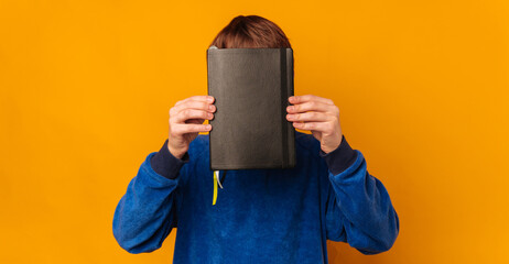 Teen boy covers his face with a black leather note book he is holding. © Vulp