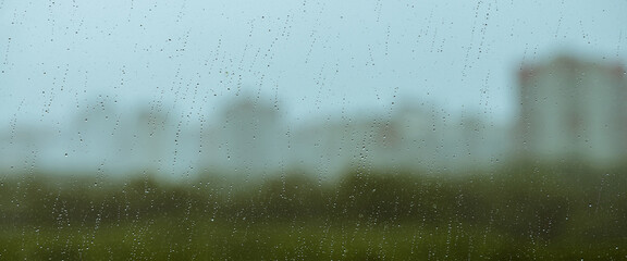 Atmospheric minimal backdrop with rain droplets on glass. Wet window with rainy drops and dirt spots closeup. Blurry buildings and green trees under blue sky against dirty window glass with raindrops.