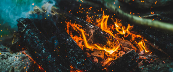 Vivid smoldered firewoods burned in fire close-up. Atmospheric warm background with orange flame of campfire and blue smoke. Full frame image of bonfire. Beautiful whirlwind of embers and ashes in air