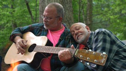 Closeup of two gay men sharing love in front of a campfire and playing guitar in a forest.