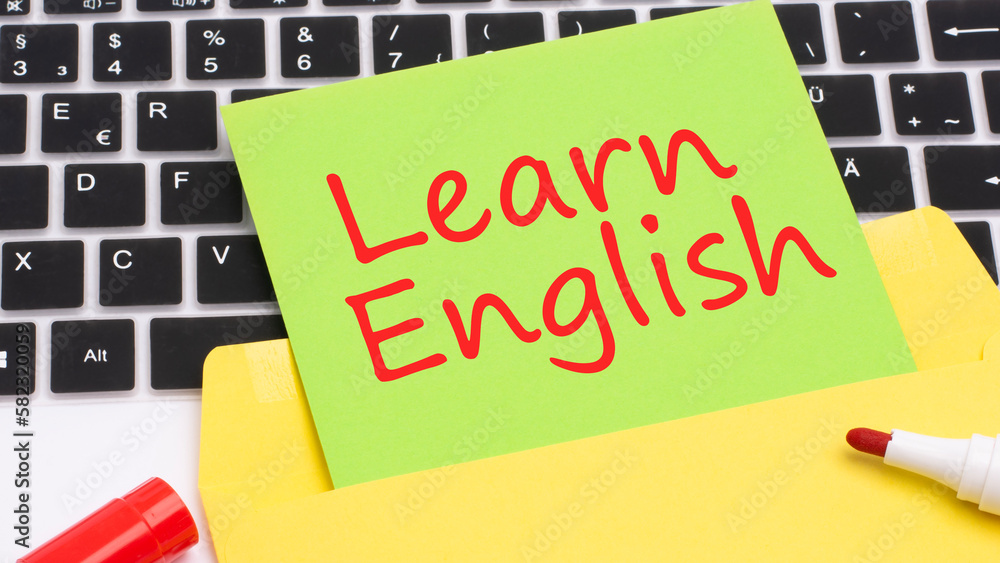 Poster text learn english is written in red marker on a green card. in the frame is a laptop keyboard on ye