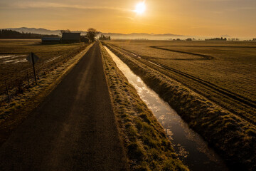 Fototapeta na wymiar Rural Country Road and Irrigation Ditch at Sunrise. Atmospheric aerial view of an historic barn and surrounding farmland.