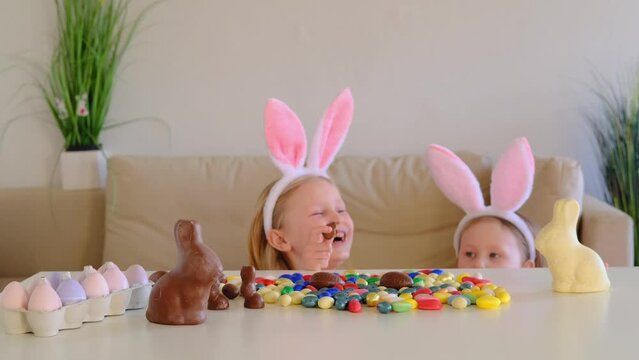 Small children, sisters with rabbit ears hunt for chocolate eggs eat them and laugh.