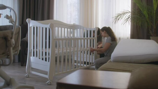 Loving parents together assembling white crib for newborn baby at nursery. Young father uses hex key for fixing mounts. Caring wife helps beloved man. Concept of parenthood, childhood and family.