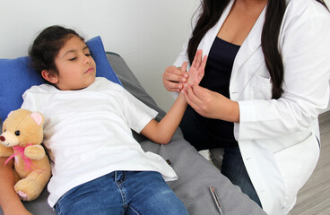 Latina female pediatrician doctor attends to her 7-year-old girl patient, checks her fingers,...
