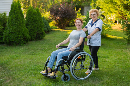 An elderly female nurse walks with a middle-aged woman in a wheelchair in the park. 