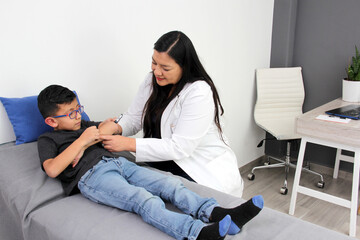 Latina female pediatrician doctor attends to her 6-year-old boy patient, checks her fingers, hands,...