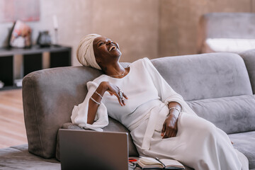 Excited African young woman in white turban and traditional dress relaxing on couch at home laughing out loud after friends joke during distant video call using laptop. Happy people, business, finance - Powered by Adobe