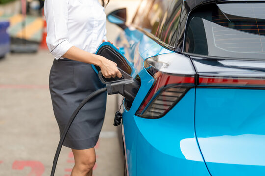 Woman plugging in the charger into a socket of her blue electric car at a charging station in the street