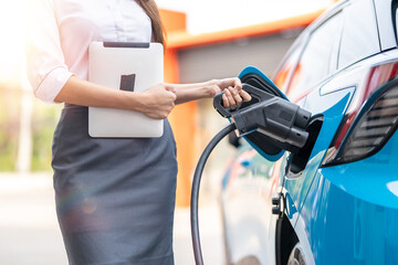 Woman plugging in the charger into a socket of her blue electric car at a charging station in the...