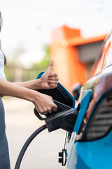 Woman plugging in the charger into a socket of her blue electric car at a charging station in the...