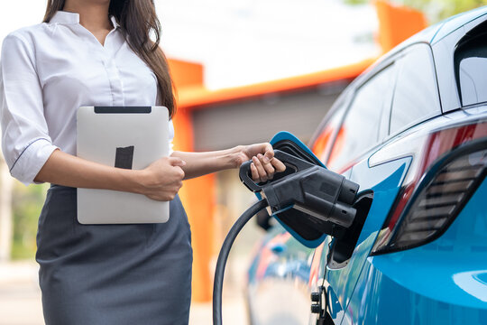 Woman plugging in the charger into a socket of her blue electric car at a charging station in the street