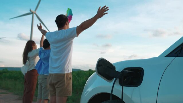 Concept of progressive happy family enjoying their time at wind farm with electric vehicle. Electric vehicle driven by clean renewable energy from wind turbine generator for charging station.