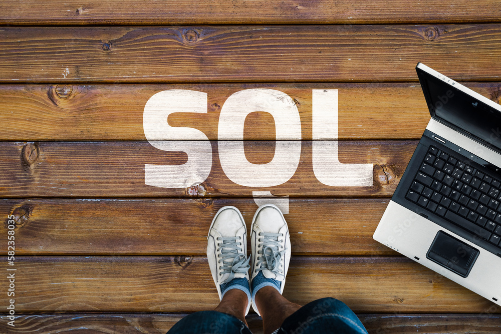Poster legs in sneakers standing next to laptop and sql on wooden floor. sql programming language