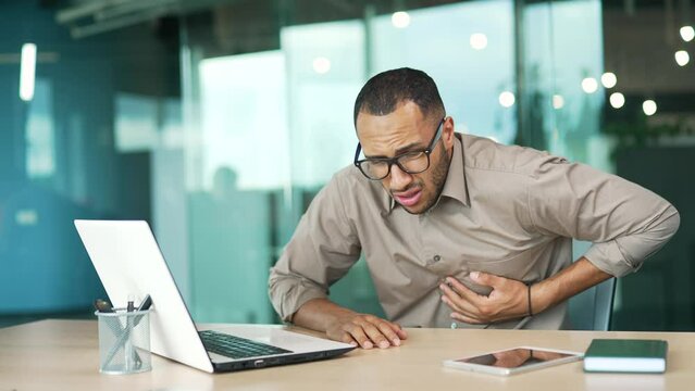 A young adult man has a heart attack while sitting at a workplace at a desk in a modern office. A sick employee of mixed race suffers from tension in the chest and holds the sore spot with his hands