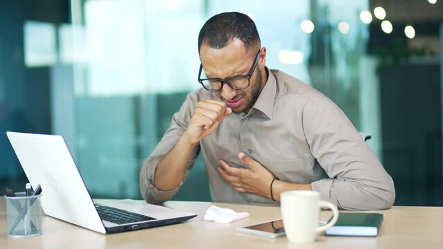 Young adult man coughing while sitting at workplace at desk in modern office. Sick with a cold or flu mixed race employee feels bad, holds his hand to his chest. He has a high temperature, bronchitis