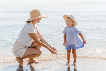 Mom with a toddler daughter in straw hats on the beach by the sea