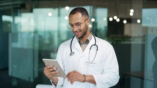 Portrait of a young smiling doctor using a tablet while standing in a clinic. Handsome mixed race therapist in glasses and white coat looks at news on the Internet, writes back messages, reads email