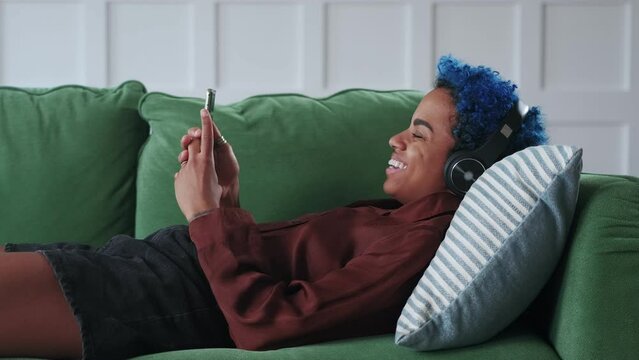 Young sociable African American woman hipster say goodbye after videocall on phone and laugh rejoicing at long-awaited Internet call from old friend lying on sofa in living room. Zoomers lifestyle