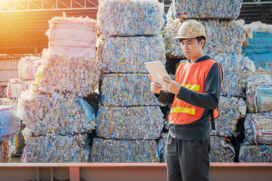 Asian engineer wearing hard hat and protective vest checking data with tablet at stack of recycled water bottles in the recycling industry, Recycling and reuse concept.