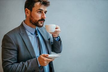 A pensive businessman standing and drinking a coffee against the gray background. Copy space