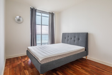 Compact minimalistic cozy double bed in bedroom with wooden laminate flooring and white walls and gray curtains with window with stunning views on sunny summer day. Hotel room concept