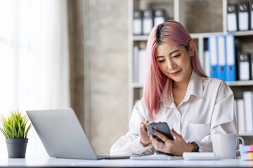 Portrait of a happy Asian businesswoman working on a laptop computer at her home office while using a mobile phone. Smiling businesswoman contacting the business to talk about online products.