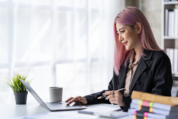 Portrait of a young Asian businesswoman sitting at a desk in an office recording data on a laptop....