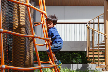 happy asian boy play on playground. boy kid climbing up on ladder at the public playground in the city park on weekend.