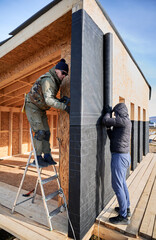 Male builders installing waterproof membrane on the wall of future cottage. Men workers building wooden frame house in the Scandinavian style barnhouse. Carpentry and construction concept.