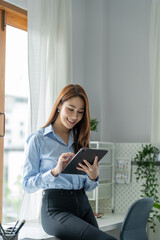 Fototapeta premium A pretty smiling Asian businesswoman holding a tablet to look at financial details, and online delivery orders in the office happily.