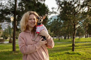 a small dog in nature with the owner in the park, a pet in the hands of a woman in dog clothes in the summer for a walk