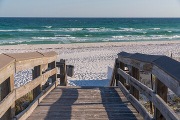 Fototapeta premium Wooden walkway with view of sea and blue sky on a sunny day in Destin Florida. The pathway leads to the sunlit shore and ocean with view of the horizon in the distance.