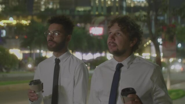 Medium shot of two multiethnic businessmen in formalwear walking along city street at night, holding to go coffee cups and having discussion afterwork