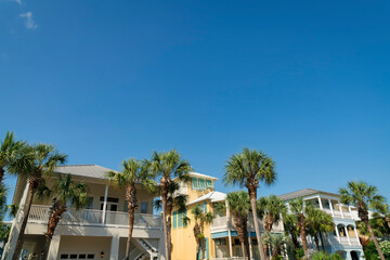 Three houses with palm trees outdoors under the blue sky at Destin, FL. There is a house left with white exterior and stairs outdoor, near the yellow house middle beside the blue house right.
