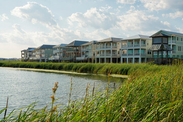 Row of houses under the bright sky with clouds with lagoon waterfront at Destin, Florida. There is a lagoon at the front with tall grasses near the shore and a boardwalk pavilion on the right. - obrazy, fototapety, plakaty
