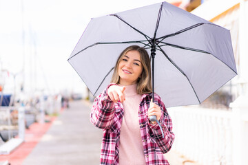 Young pretty Romanian woman holding an umbrella at outdoors points finger at you with a confident expression