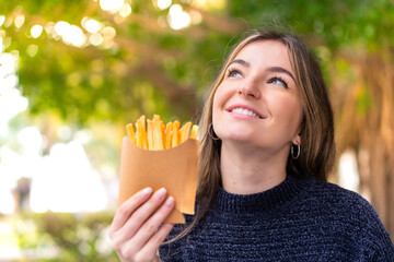 Young pretty Romanian woman holding fried chips at outdoors looking up while smiling