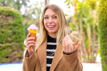 Young pretty blonde woman with a cornet ice cream at outdoors inviting to come with hand. Happy...