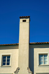 Fototapeta na wymiar Tall chimney with metal vents and dark adobe roof tiles with visible windows with white frames and beige stucco cement