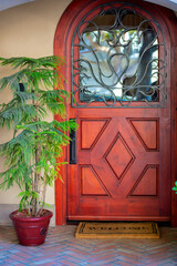Wooden front door with decorative facade and glass windows with brown welcome mat and tropical plant in pot