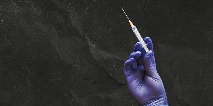 Gloved hand of a doctor or nurse holding a syringe with liquid vaccine before injecting over dark background with copy space on the right.