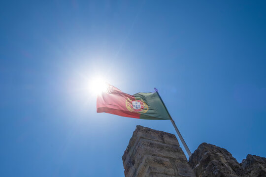 the national flag of portugal flies on top of a battlement of the castle of St. George in Lisbon on a sunny day, blue sky, horizontal